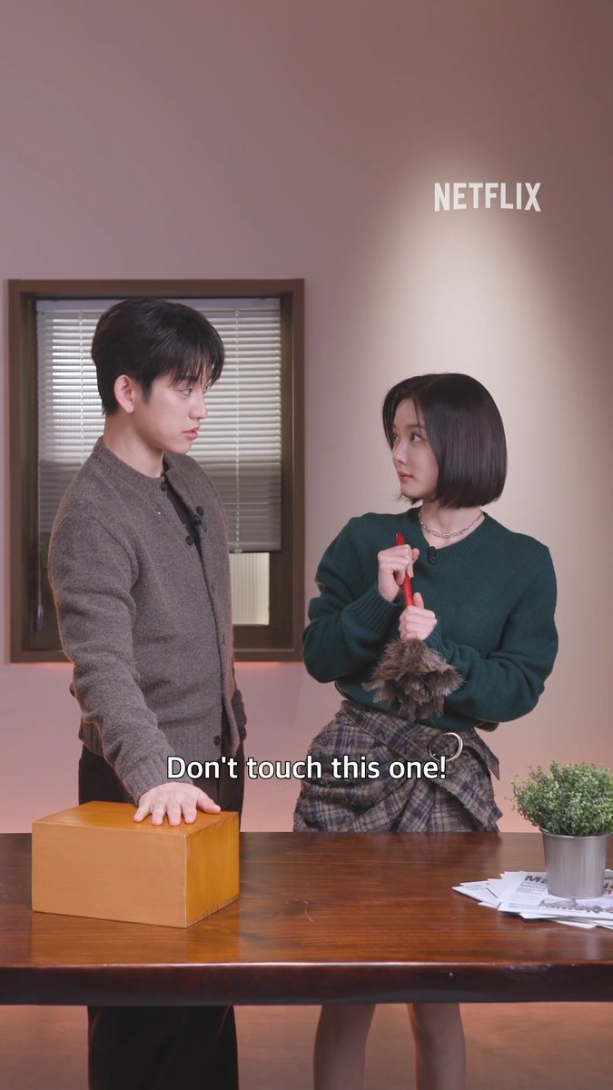 A Netflix promotional image showing two people—a young man in a gray cardigan and a young woman with a bob haircut holding a red pen—standing near a table with a wooden box, with the subtitle 'Don't touch this one!'