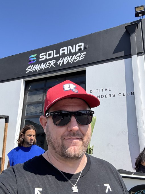 A man wearing a red Solana-branded cap, sunglasses, and a black t-shirt takes a selfie in front of a building with a 'SOLANA SUMMER HOUSE' sign.