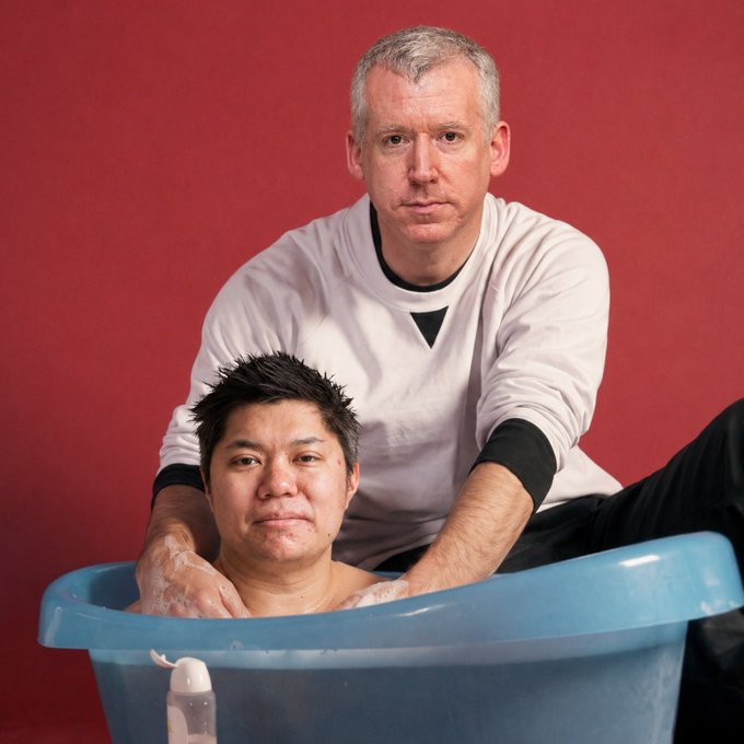 Two men, one in a white sweatshirt, in a bathtub with a red backdrop in what appears to be a promotional or comedic photo.