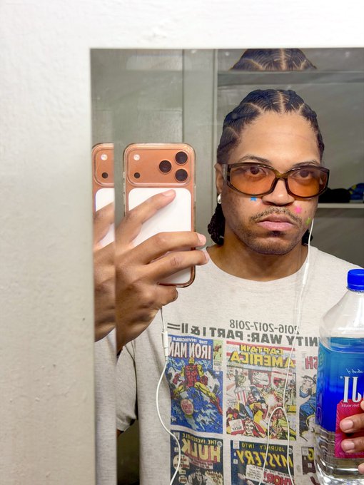 A man with braided hair and orange-tinted sunglasses takes a mirror selfie while holding a bottle of Fiji water, wearing a vintage comic book-printed t-shirt featuring titles like "Iron Man," "Avengers," and "Hulk," along with "Nitty War Part 12!"—fitting perfectly with the caption "The Fiji is part of the fit."