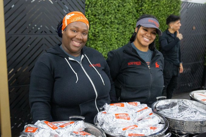 The image shows two women standing next to each other behind a table filled with food items such as sandwiches and chips. They are smiling and appear to be enjoying their time together, possibly working at an event or gathering where breakfast is being served. The background features a green wall that adds color to the scene. There is also another person visible in the image who seems to be engaged in conversation or observing something else.