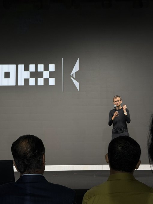 A man giving a presentation on stage in front of a large 'OKX' logo projection, addressing an audience visible in the foreground.