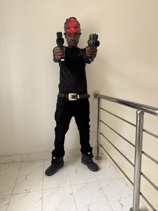 A young person poses in an all-black outfit, accessorized with a distinctive red demon-like mask and a leopard-print belt, dual-wielding what appear to be prop guns while standing near a metal railing. The edgy, action-themed aesthetic complements the post's energetic "gm 𝕏 new week, let's get it" message, projecting a fierce, motivational vibe to kick off the week.