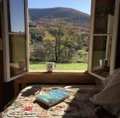 A view through an open window onto a rural landscape with rolling hills and a winding dirt road, with a book resting on a cushioned window seat.