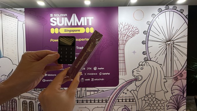 A person holds a small black device (likely a SafePal hardware wallet) and a branded SafePal snack bar in front of a vibrant purple "Solana Summit Singapore" backdrop featuring the Merlion and Singapore Flyer. The image showcases the SafePal booth setup at the event, aligning with the post about being ready to connect with attendees at the Ritz Carlton Grand Ballroom.