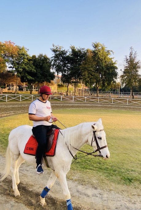 A rider wearing a red helmet and white "CRONA" shirt sits on a white horse in a fenced outdoor arena, with "CRONA" also visible on the red saddle pad. The image fits the cheerful weekend greeting, capturing a relaxed morning ride as the rider likely sends out their "GM" (good morning) message to the community.