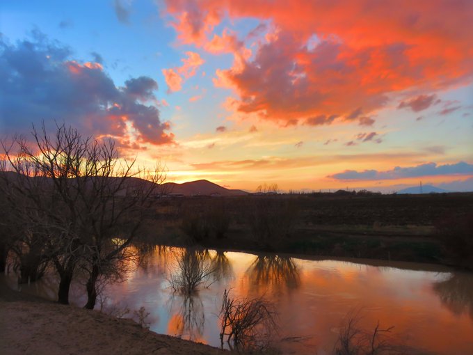 A scenic sunset over a tranquil pond, with vibrant orange and pink clouds reflecting on the water's surface, framed by silhouetted bare trees.