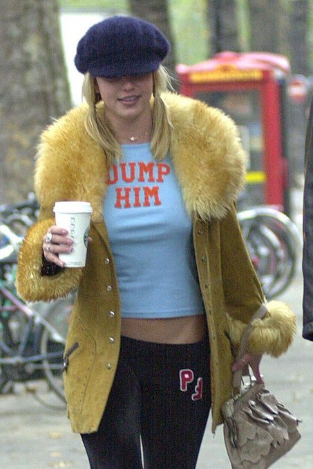Britney Spears walking outdoors wearing a blue beret, a yellow fur-collared jacket over a "DUMP HIM" crop top, holding a coffee cup and a handbag.