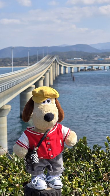 Photo of a stuffed dog toy wearing a yellow cap, red varsity vest, and a small camera, posed in front of a long bridge spanning the sea.