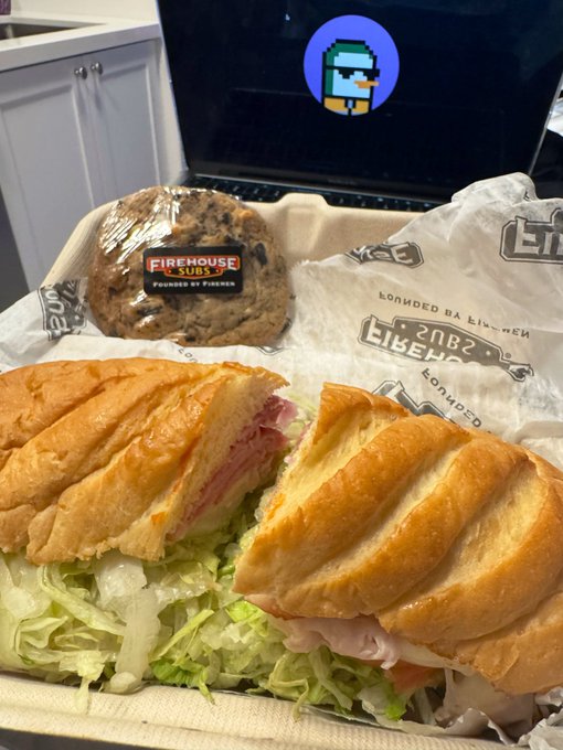 A Firehouse Subs meal featuring a sliced sandwich overflowing with lettuce and meat, accompanied by a chocolate chip cookie with "FIREHOUSE SUBS" branding on the wrapper. The caption "time to make more happen" suggests a motivation to fuel up and tackle productivity with a hearty sub from the well-known chain.