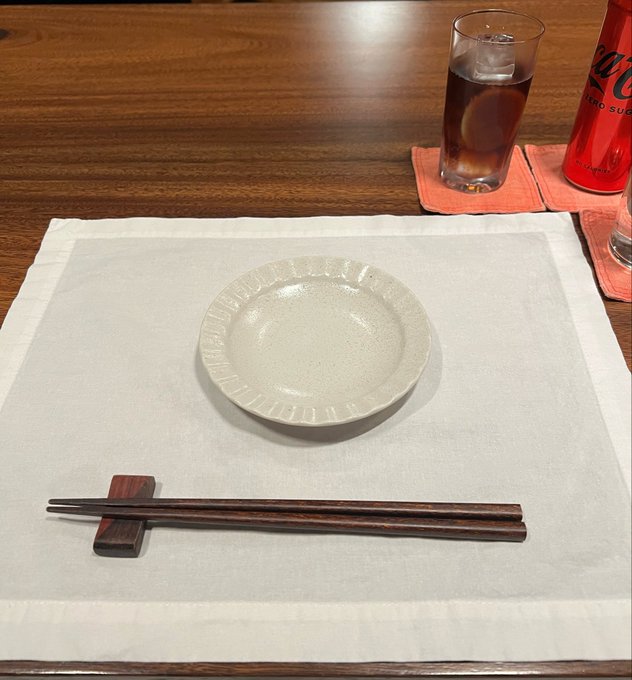 A place setting featuring a small cream-colored plate resting on a white placemat, with dark wooden chopsticks on a chopstick rest and a glass of iced beverage nearby.