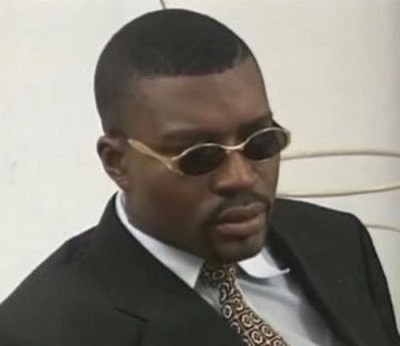 The image features a man wearing sunglasses and a suit jacket sitting down with his head tilted back, looking off into the distance. The background of the photo appears to be white, which contrasts with the man's dark hair and the black suit he is wearing. There are no texts or other objects in the image, making it a simple yet striking composition that captures the viewer's attention.