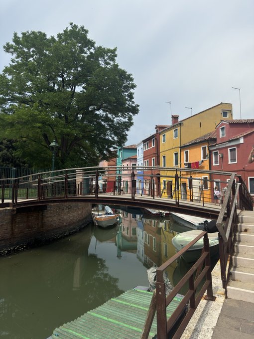 A picturesque canal scene featuring a pedestrian bridge, small boats moored along the water, and colorful buildings in shades of yellow, orange, and red.