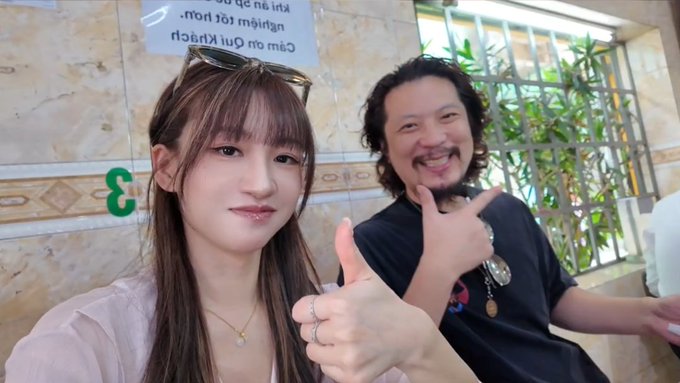 Two people taking a photo together, both giving thumbs up, with a sign in the background featuring mirrored Thai text.