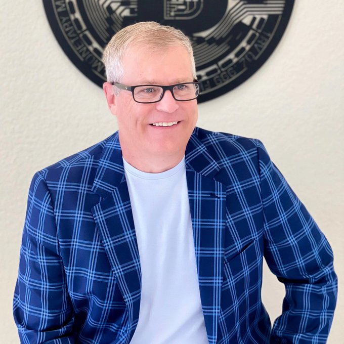 A smiling man with glasses wearing a blue plaid blazer over a white shirt, posing in front of a seal that reads 'UNITED STATES NAVY MONEY MATTERS'.