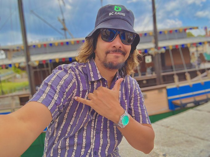 A man wearing a "BC.GAME" bucket hat and sunglasses flashes a shaka sign while sporting a purple patterned shirt and green-faced watch, with colorful nautical flags and boats visible behind him, channeling relaxed "Gm Fam" vibes for the crypto community.