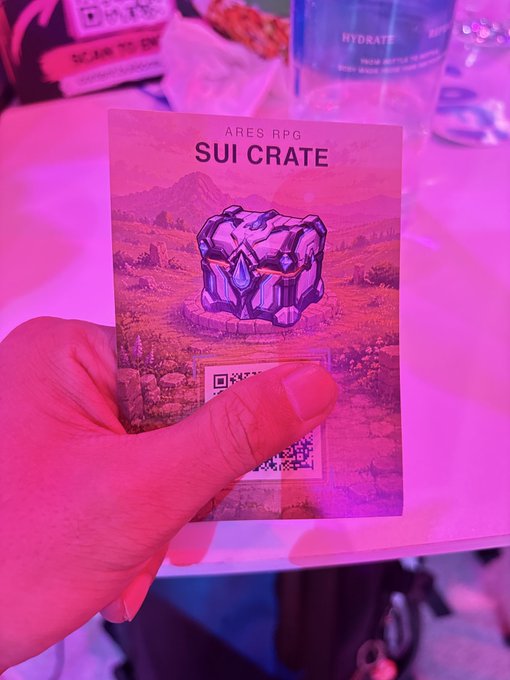 A hand holds a card labeled 'ARES RPG SUI CRATE' featuring an illustrated treasure chest with a QR code, in pink-lit surroundings.