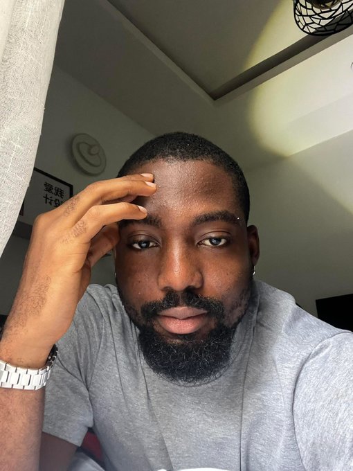 A selfie of a man with a dark beard wearing a grey t-shirt, resting his hand on his forehead in a softly lit room.