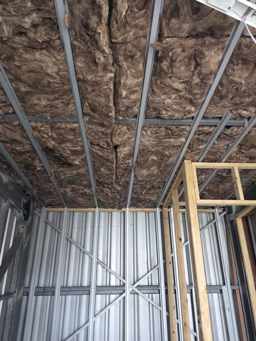 The interior of a building under construction showing exposed steel ceiling framing filled with brown rockwool insulation above corrugated metal walls.