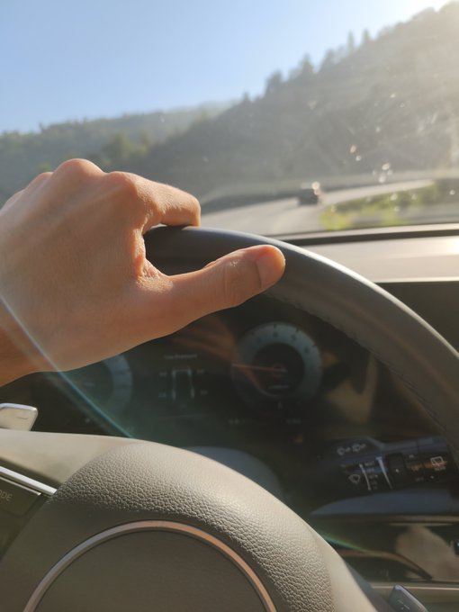 A close-up view from the passenger seat of a person's hand gripping a car steering wheel while driving on a sunny road with hills in the background.