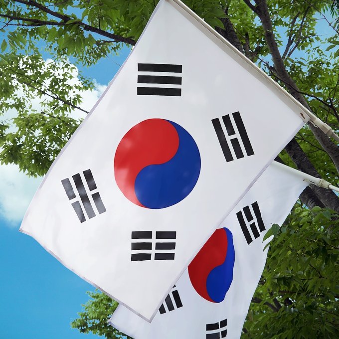 Two South Korean flags hang against a backdrop of green tree branches and blue sky, symbolizing the nation where police have reportedly booked 26 Polymarket users for illegal gambling and referred 18 to prosecutors over $12.7M in combined wagers.