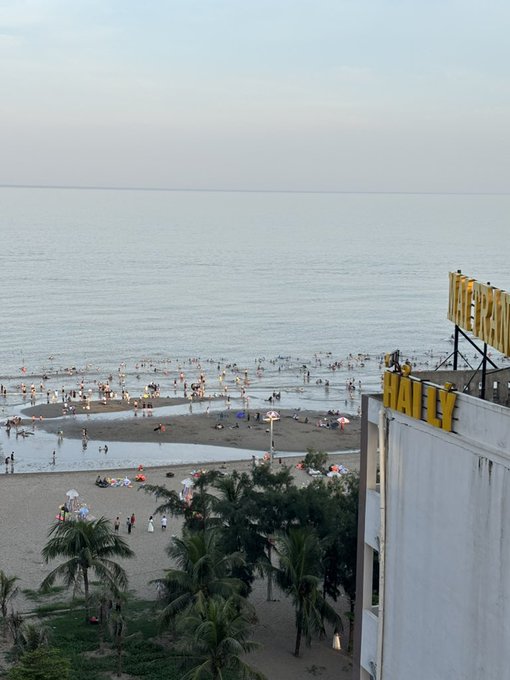 An aerial view of a crowded beach with people swimming and wading in the ocean, with palm trees in the foreground and a yellow hotel building partially visible on the right side.