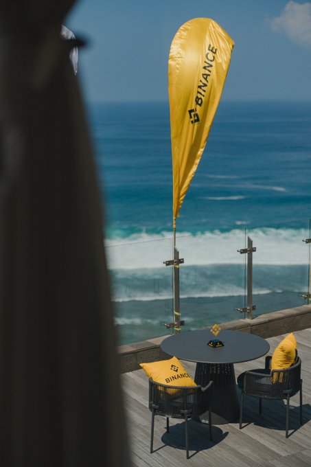 A bright yellow Binance feather flag sways over an oceanfront terrace featuring a small bistro table flanked by two chairs adorned with "BINANCE" branded cushions, with only one cushion visible from this angle—perfectly matching the post's invitation to claim your seat and bring a plus one.