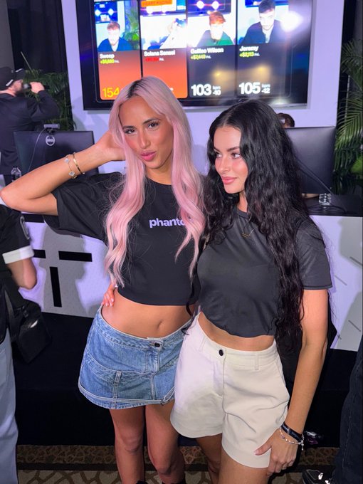 Two women, one with pink hair wearing a "phantom" crop top and denim skirt and another with long dark hair in a black top and white shorts, pose together at what appears to be a stock trading event, with screens behind them displaying tickers like "DUKC" at $154, "103", and "105" — humorously captioned "this is who you're trading against."