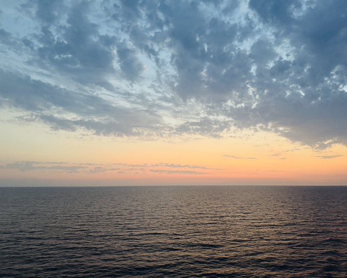 Serene photograph of an ocean seascape at sunset or sunrise, featuring a calm dark water surface and a sky filled with scattered clouds tinted in soft orange and blue hues.