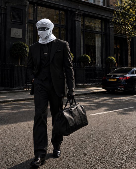 A fashion photograph of a person dressed in an all-black three-piece suit with a white face wrap and sunglasses, carrying a black leather bag on a London street near '26 LOND SQUARE'.