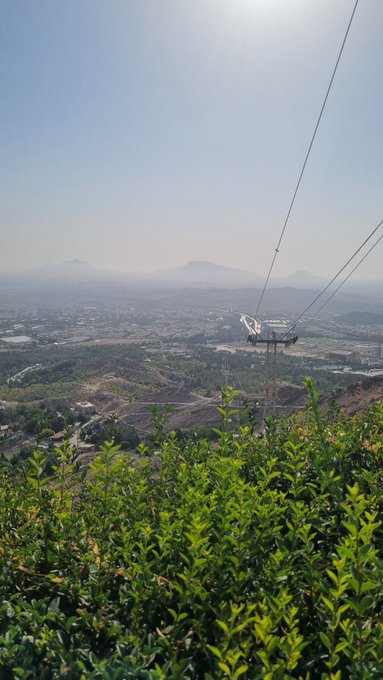 A scenic vista overlooking a sprawling city and hazy mountains from a hillside, featuring a cable car tower and power lines in the foreground.