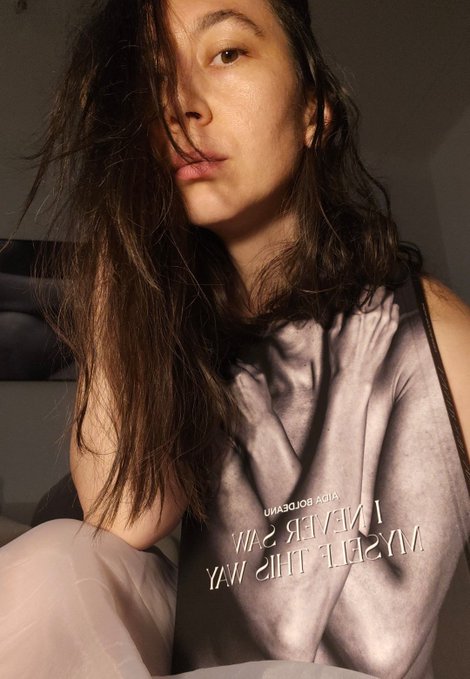 A woman with long, tousled hair poses in a sleeveless gray top featuring the visible text "KISS, THE SUN. NEVER BE SHY." The intimate, softly lit portrait complements the celebratory post about being the final person to mint a sold-out art book, capturing the personal excitement of the collector.