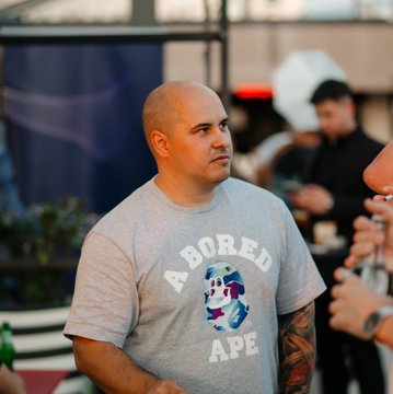 A man wearing a gray "Bored Ape" t-shirt stands in what appears to be an event setting.