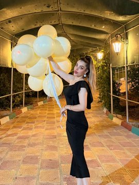 A woman with a high ponytail wearing a black crop top and matching mermaid-style skirt holding a large bunch of cream-colored balloons in a covered walkway at night.