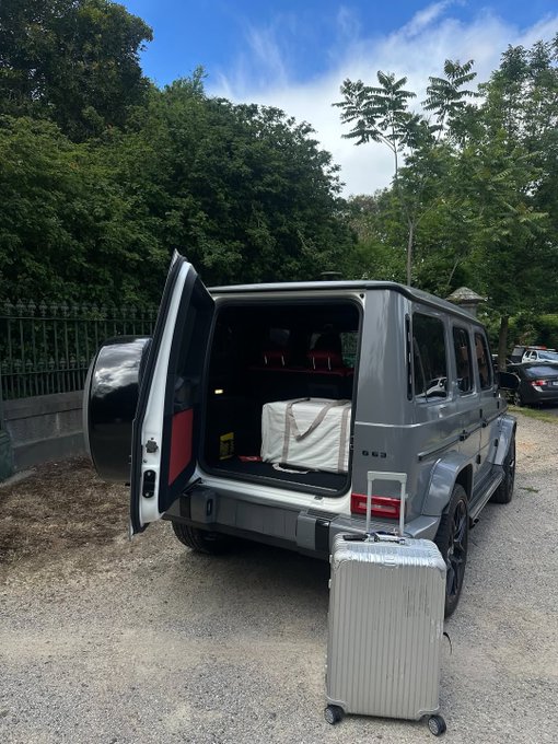 The image shows a silver SUV with its trunk open, revealing two suitcases inside. The trunk of the car is filled with luggage, indicating that someone might be preparing for a trip or has recently returned from one. There are also other cars visible in the background, suggesting that this scene takes place on a busy street or parking lot.