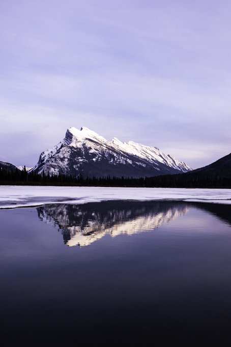 A snow-capped mountain is reflected in the calm waters of a partially frozen lake under a soft purple and blue sky at dusk or dawn.