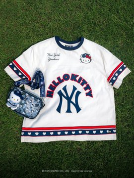A white Hello Kitty New York Yankees collaboration t-shirt lies on grass next to a matching blue Hello Kitty handbag.