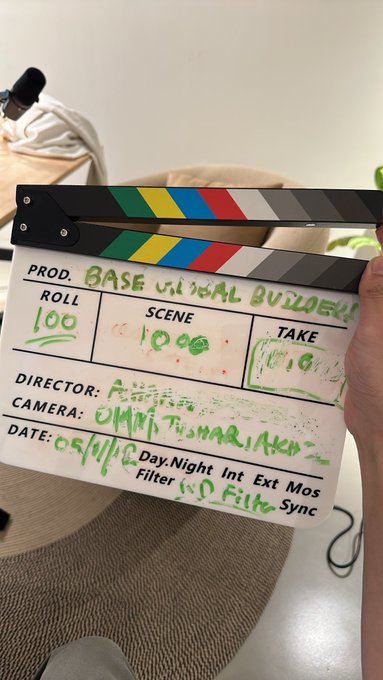 A hand holds up a clapperboard labeled "BASE MORAL BUILDERS" in green marker, with Roll 100, Scene 10, Take 01, Director "P..." (partially illegible), Camera "Omni Tuomarilaw" (partially illegible), and dated 05/16, capturing a candid behind-the-scenes moment on a film or video set.