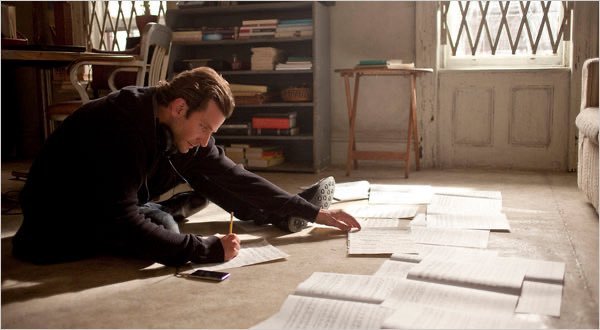 A man with earphones sits on the floor of a vintage room, writing on papers spread across the floor with a pencil.