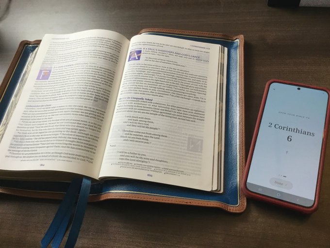 An open Bible in a leather cover rests on a wooden table next to a smartphone displaying '2 Corinthians 6' on the 'OPEN YOUR BIBLE' app.