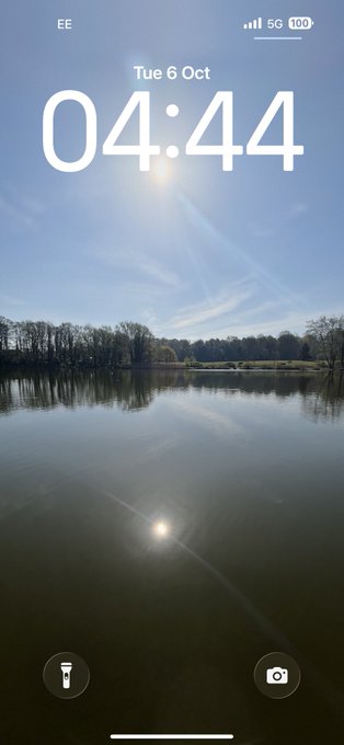 A phone lock screen displays the time '04:44' on 'Tue 6 Oct' over a background image of a calm lake reflecting a sunlit sky and trees.