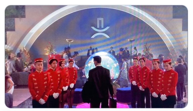 A scene from a movie or show featuring red-uniformed attendants lined up symmetrically in front of a glowing globe, with a person in a black suit walking toward it under an archway displaying a logo.