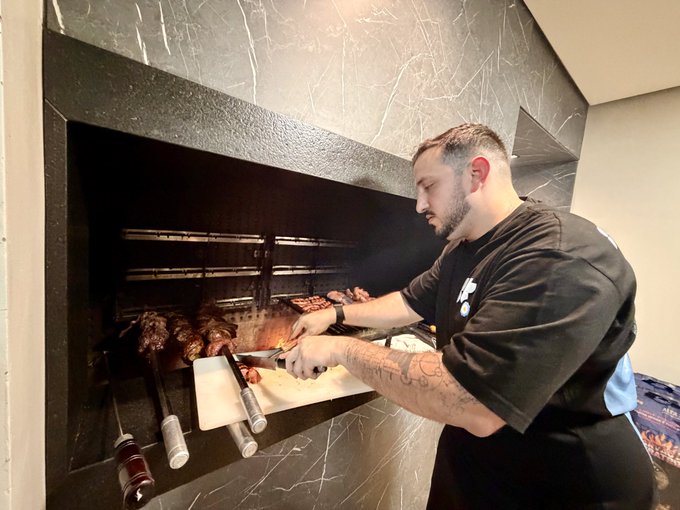 A man in a black shirt grills skewers and meat over an open flame in a modern indoor kitchen. The caption “Im cooking at @islanddao not joking” suggests he is cooking at IslandDAO’s venue; no other clearly readable text or numbers are visible.