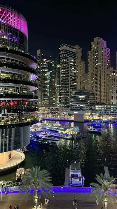 A vibrant nighttime cityscape showing illuminated high-rise buildings, docked yachts, and palm trees along a marina.