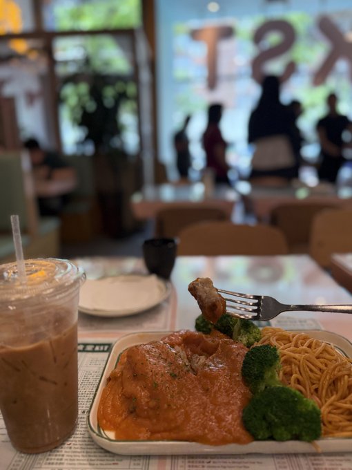 A plate of food featuring meat in red sauce with broccoli and spaghetti, accompanied by an iced coffee, photographed at a restaurant table with an 'XS' sign visible through the window.