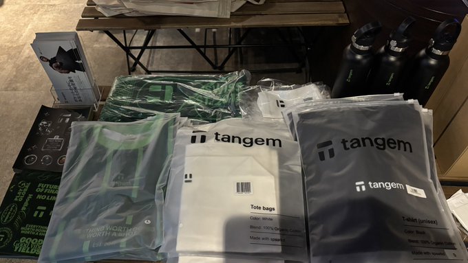 An assortment of Tangem-branded merchandise is spread on a table, including tote bags, a T-shirt, and clothing packaging, alongside black water bottles and a postcard featuring a cat.