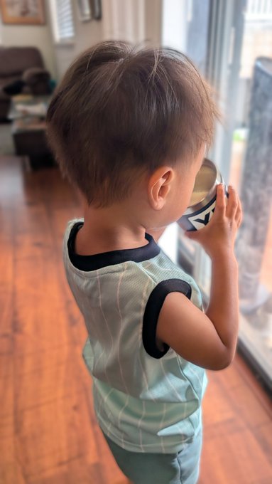 A young boy in a mint green tank top stands in a living room, sipping from a small cup containing milk. The image accompanies a "GM web3 ohana" morning greeting, with the boy's milk serving as a wholesome, kid-friendly substitute for the coffee referenced in the post.