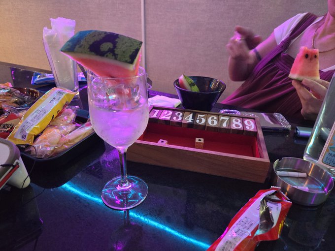 A dimly lit table scene with purple and blue neon lighting, featuring a cocktail glass with fruit garnish, a wooden box with numbered tiles 1-9, snacks, and a person in the background holding a slice of watermelon.