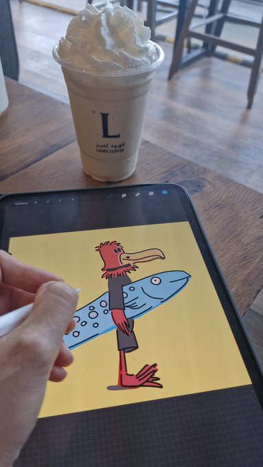 A hand using an Apple Pencil to draw on an iPad showing an illustration of a red bird-like character holding a blue fish, with an "L" branded iced coffee topped with whipped cream sitting on the table in the background.