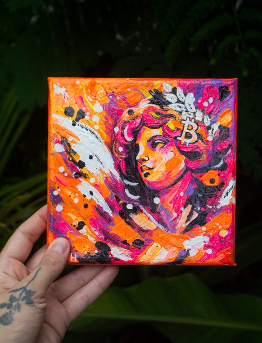 A hand holds a small, vibrant acrylic painting of a woman with pink hair and a Bitcoin 'B' symbol in her hair, painted with thick, expressive brushstrokes in bright pinks, oranges, and purples.